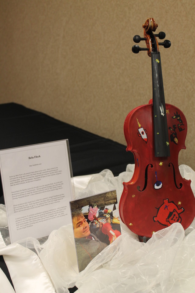 Violin painted by musician Béla Fleck. The drawings on this particular violin are to convey several things: something about our city (the rocket), the fireflies are representative of our Crescen-Dough Auction theme, and he wanted to add something about himself. The name of his second album is "Flight of the Cosmic Hippo." The drawing at the bottom right is a red hippo, which is representative of his new album "Flight of the Cosmic Hippo." We have added a link to the YouTube video: https://www.youtube.com/watch?v=1iYgf6PWYK8